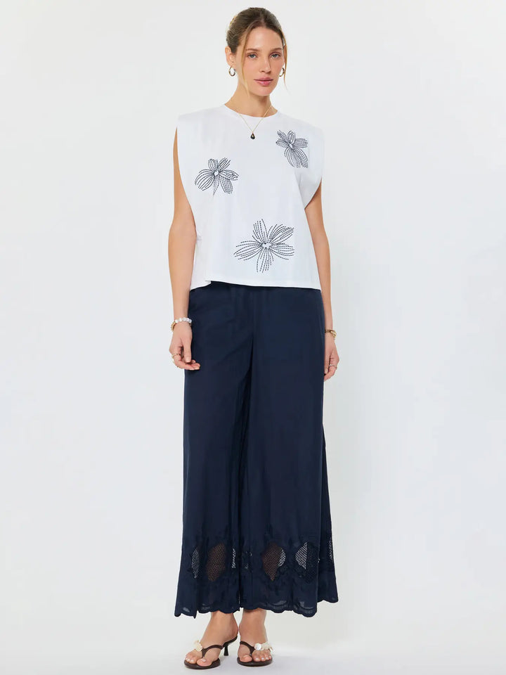 Woman wearing a white sleeveless top with floral patterns and navy wide-leg pants on a white background