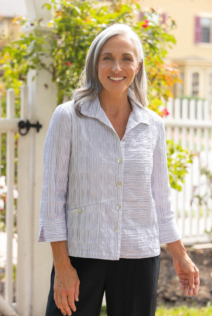 Woman wearing a light blue striped shirt standing outdoors with greenery and a white fence in the background.