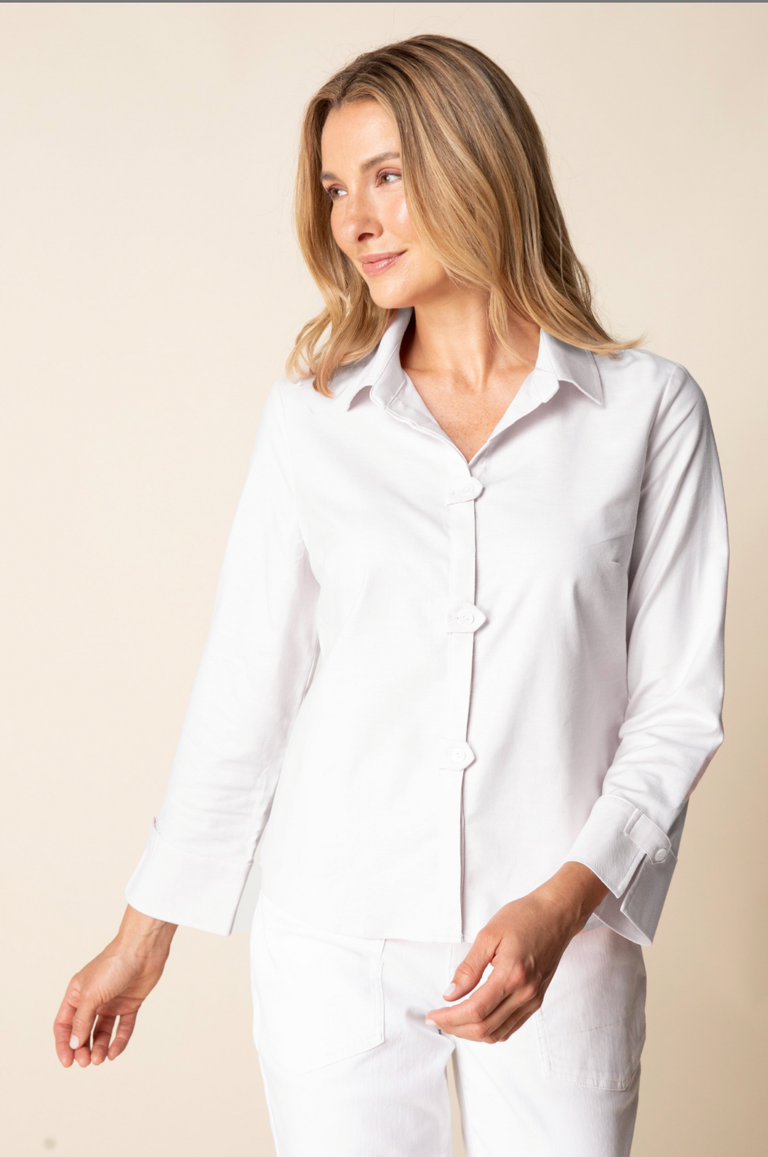 A woman wearing a white long-sleeve button-up shirt and white pants, standing against a plain beige studio background.