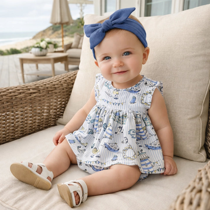 Baby wearing a patterned dress and blue headband sitting on a couch outdoors.