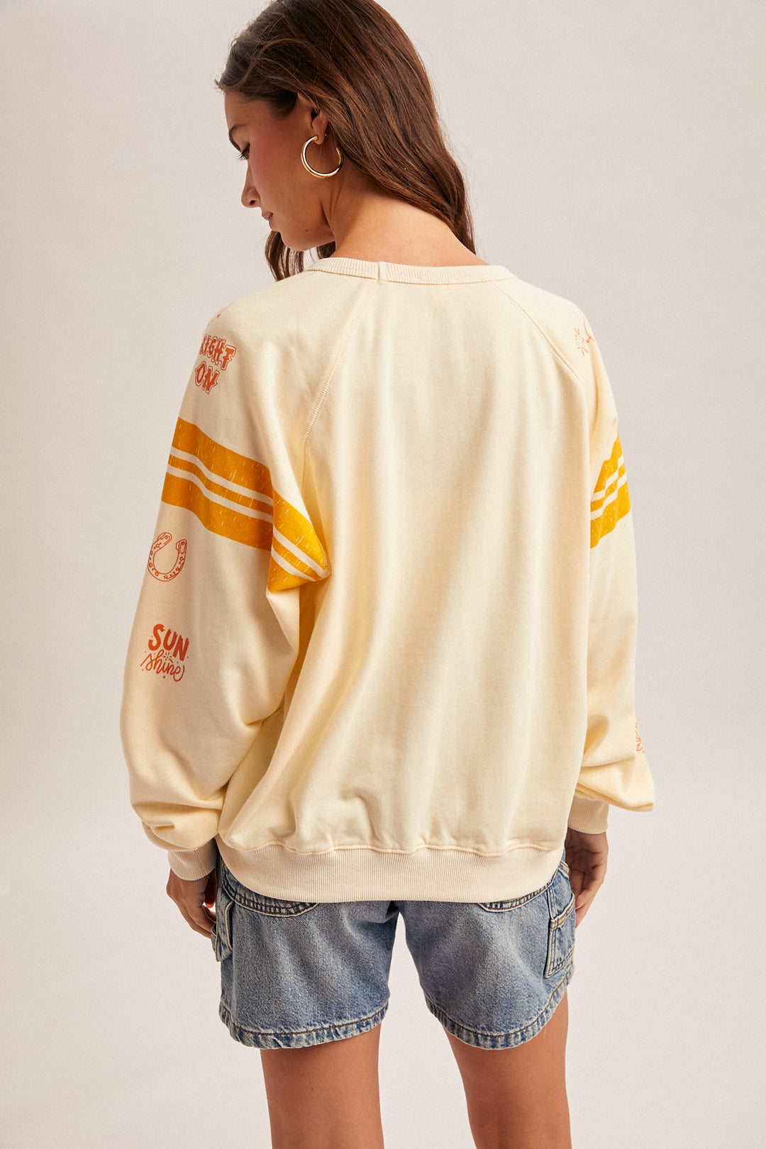 Woman wearing a cream-colored sweatshirt with yellow accents and denim shorts on a plain background