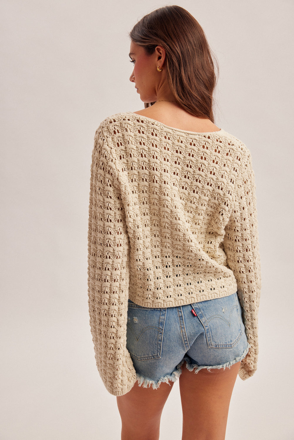 Woman wearing a beige crochet sweater and denim shorts on a plain background