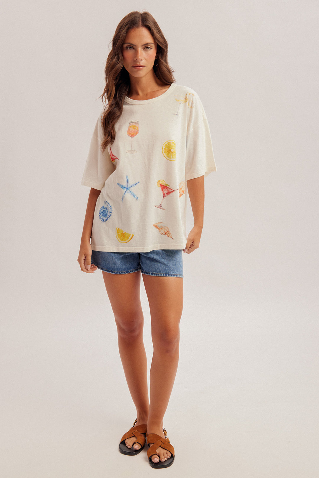Woman wearing a cream-colored t-shirt with colorful graphics and denim shorts on a plain background