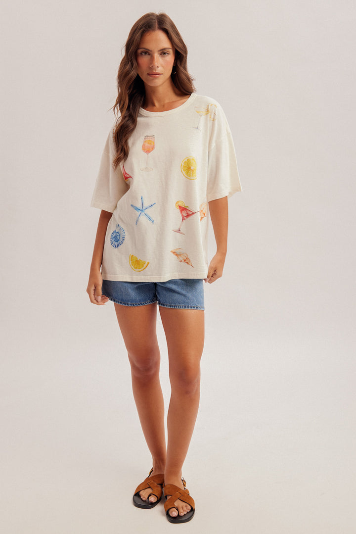 Woman wearing a cream-colored t-shirt with colorful graphics and denim shorts on a plain background