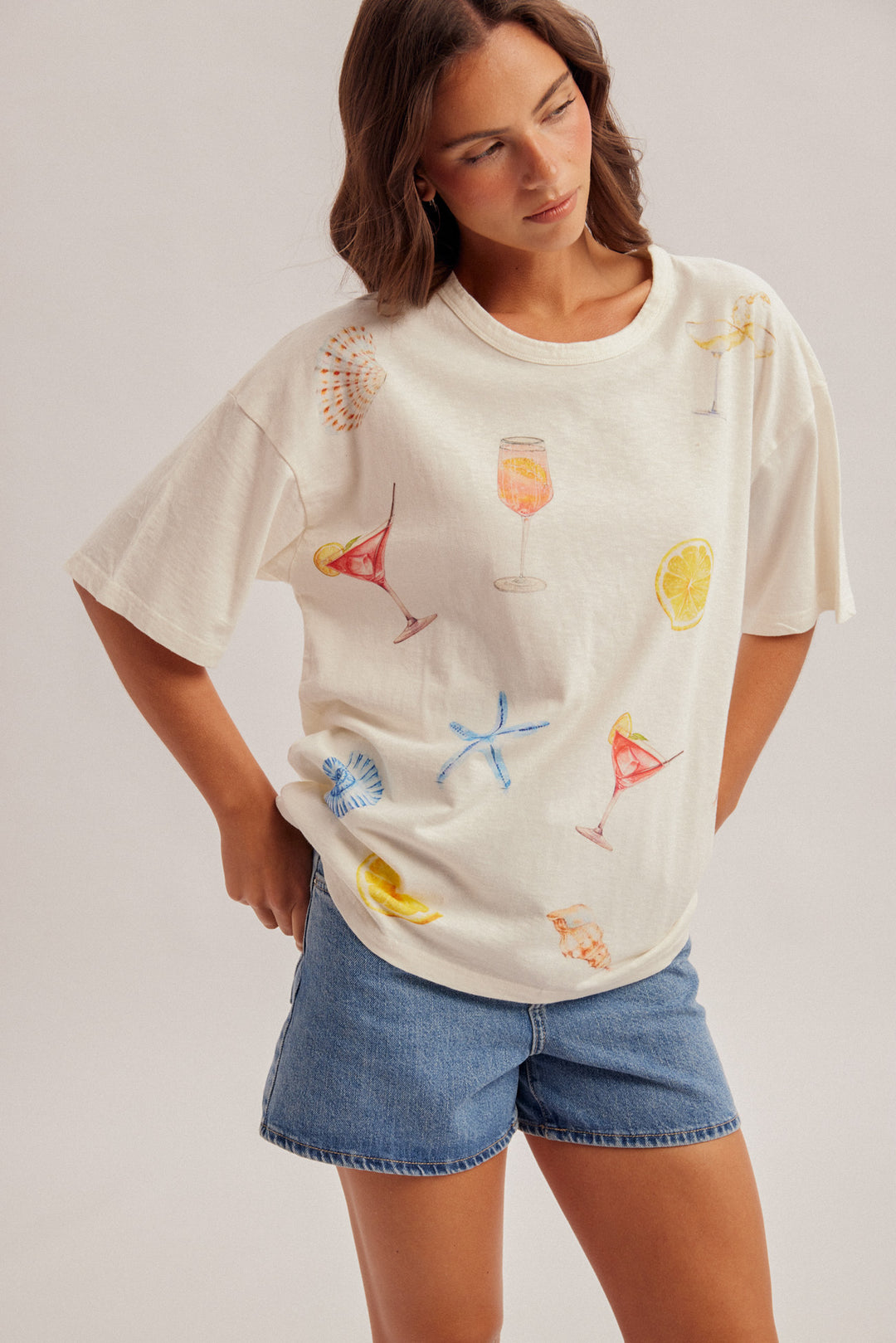 Woman wearing a white t-shirt with colorful drink graphics and denim shorts on a plain background