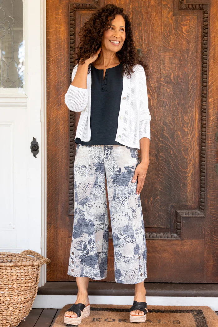 Woman standing in front of a wooden door wearing a white jacket, black top, and floral pants.