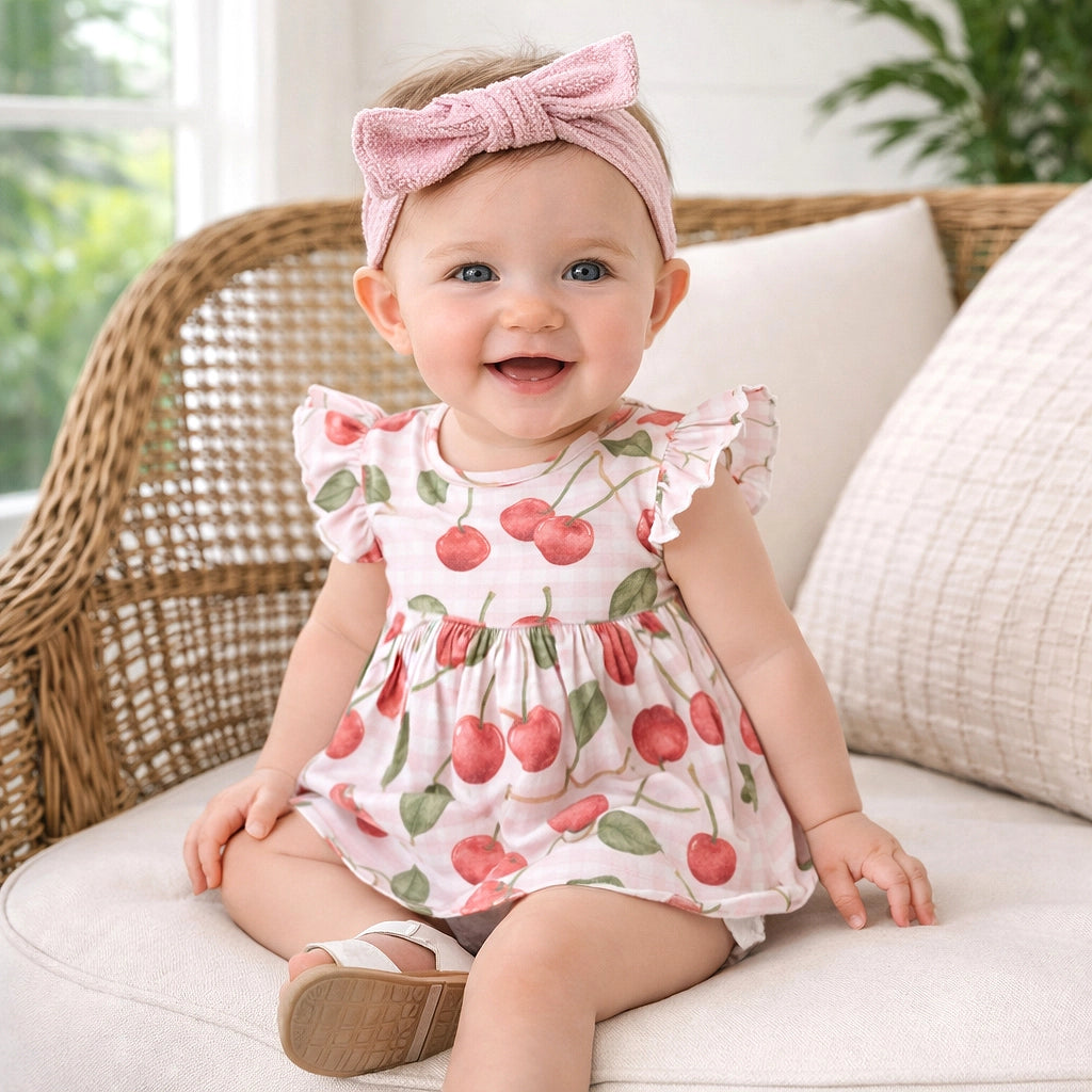 Baby wearing a cherry print dress and pink headband sitting on a couch.