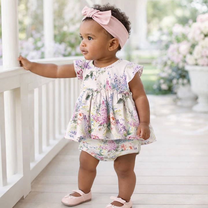 Baby girl in a floral dress standing on a porch with flowers in the background
