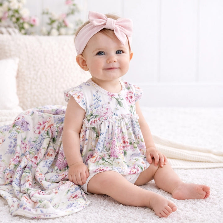 Baby in a floral dress with a pink headband sitting on a soft surface.