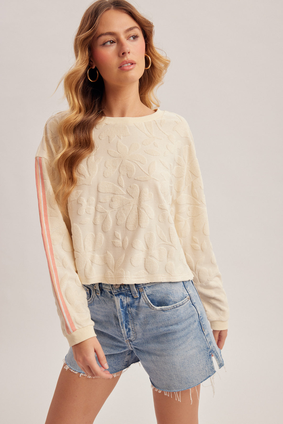 Woman wearing a cream sweater with pink stripes and denim shorts on a plain background