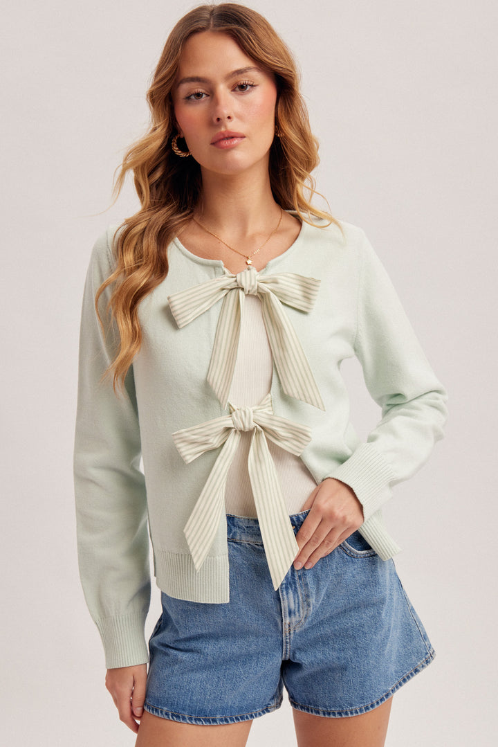 Woman wearing a light green cardigan with a bow detail and denim shorts on a plain background