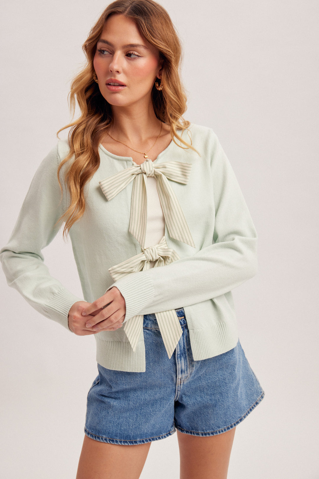 Woman wearing a light green sweater with a bow tie and denim shorts on a plain background