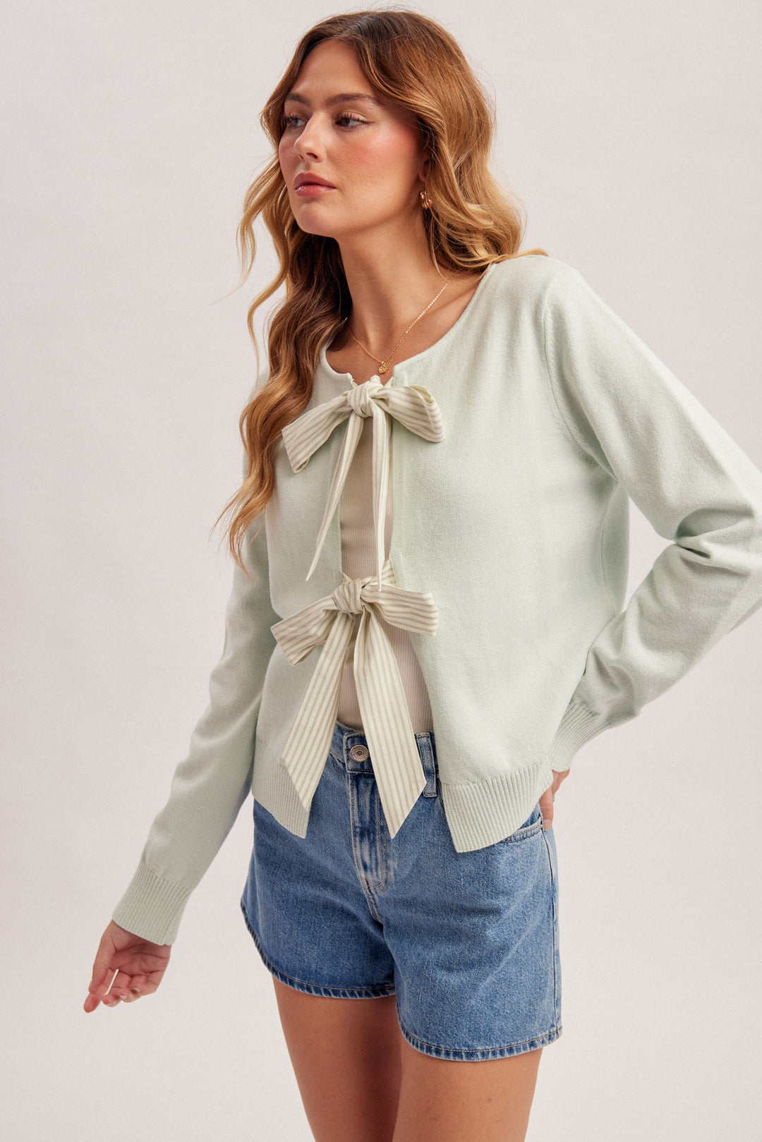 Woman wearing a light green sweater with a tie detail and denim shorts on a plain background