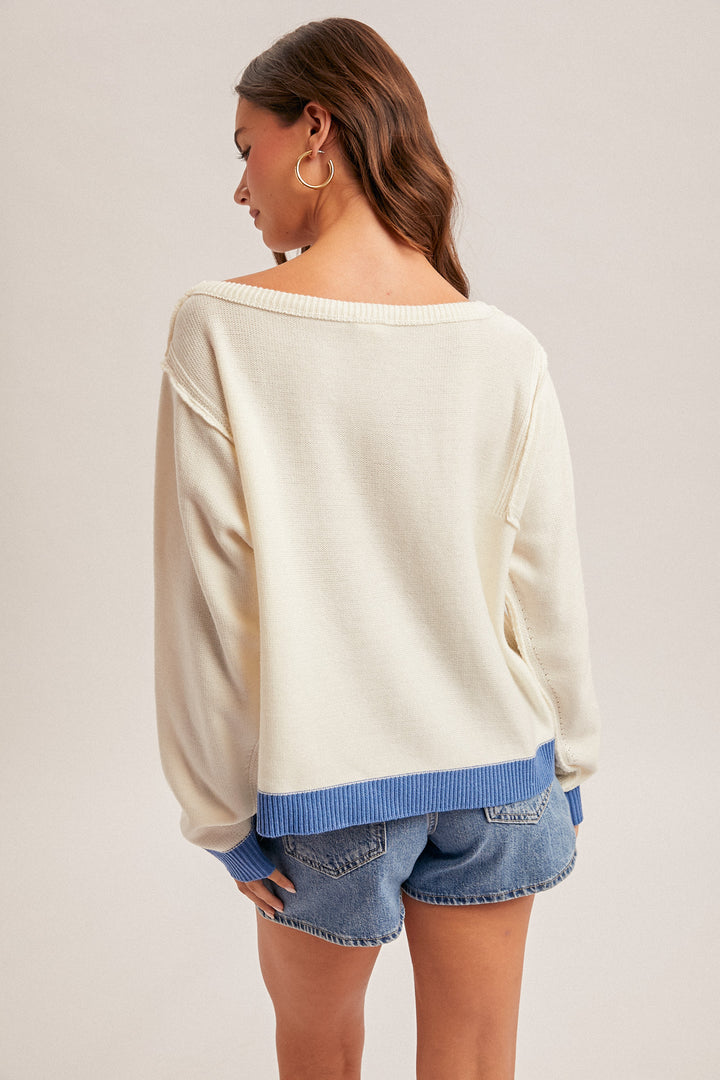 Woman wearing a cream sweater with blue trim and denim shorts on a plain background
