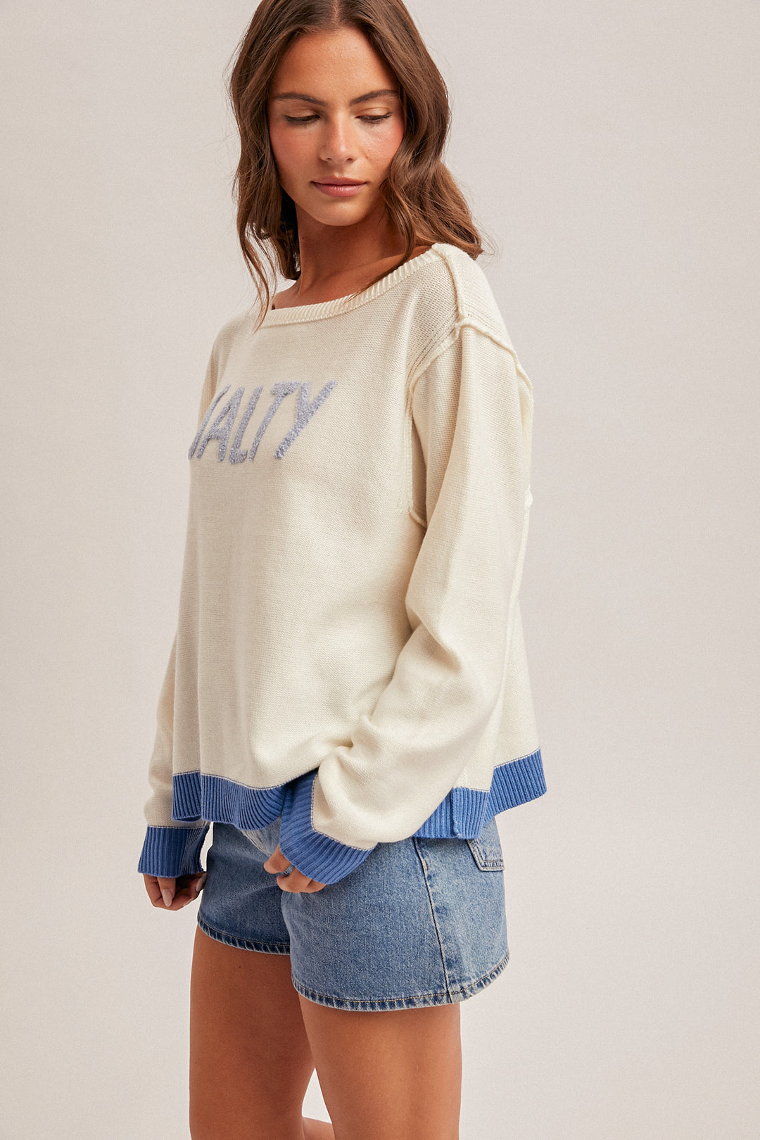 Woman wearing a cream sweater with blue text and blue cuffs and hem, paired with denim shorts on a light background.