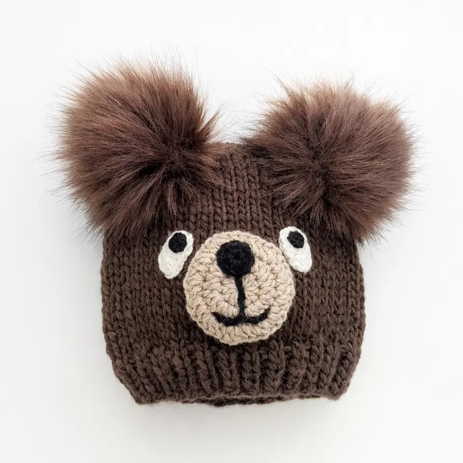 A brown knit bear-face beanie hat with two fluffy faux-fur pom-poms on top, white embroidered eyes, a beige stitched snout, and a black nose, shown on a plain white background.