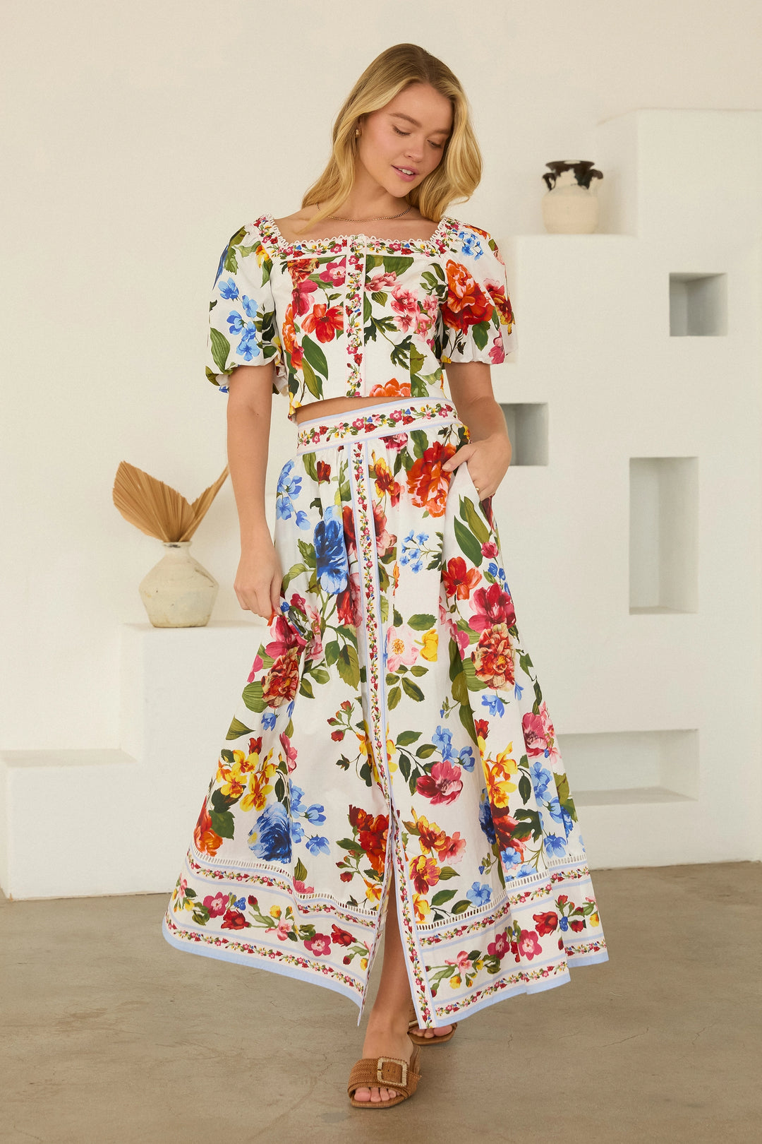 Woman wearing a colorful floral dress standing against a white wall.