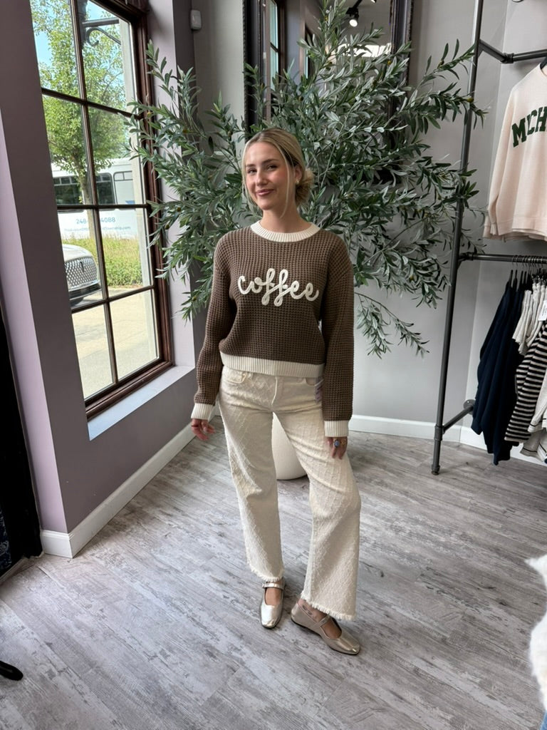 A woman stands in a boutique wearing a brown ribbed knit sweater with 'coffee' written across the chest in white script, paired with cream wide-leg pants and beige flats. A large potted olive tree and clothing racks are visible in the background.