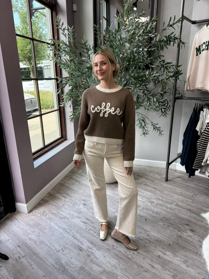 A woman stands in a boutique wearing a brown ribbed knit sweater with &