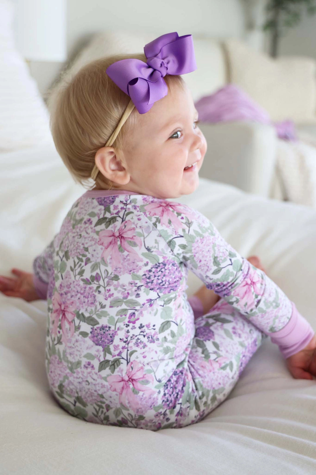 Baby in a floral onesie with a purple bow sitting on a white couch.