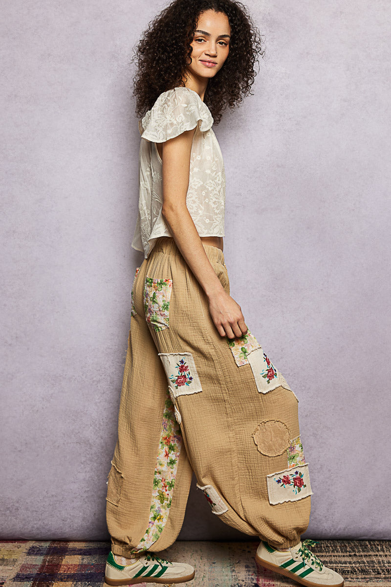 Woman wearing a white top and beige pants with floral patches against a plain background