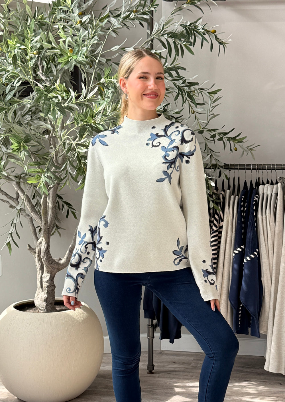 A woman modeling a cream mock-neck knit sweater with blue floral embroidery on the chest and sleeves, paired with dark blue jeans in a boutique setting.