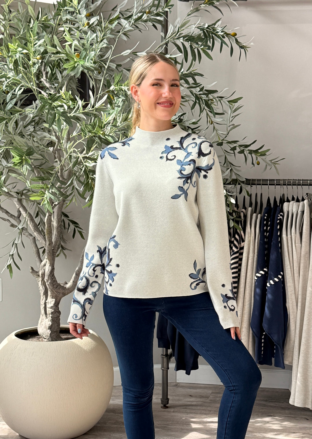A woman modeling a cream mock-neck knit sweater with blue floral embroidery on the chest and sleeves, paired with dark blue jeans in a boutique setting.