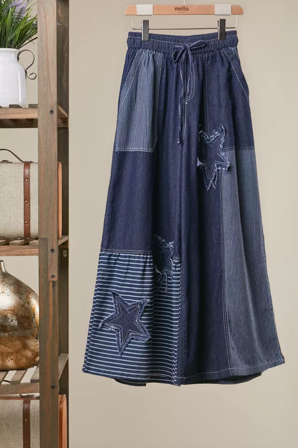 Long denim pants with star patterns hanging on a wooden hanger against a beige wall.