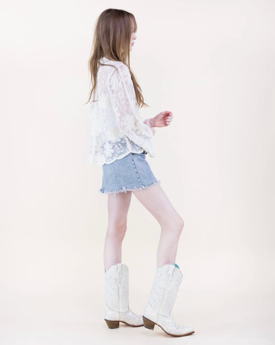 Woman wearing a beige lace top, denim shorts, and white cowboy boots on a white background