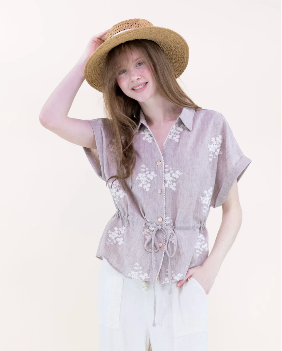 Woman wearing a light purple floral shirt and white pants with a straw hat on a plain background