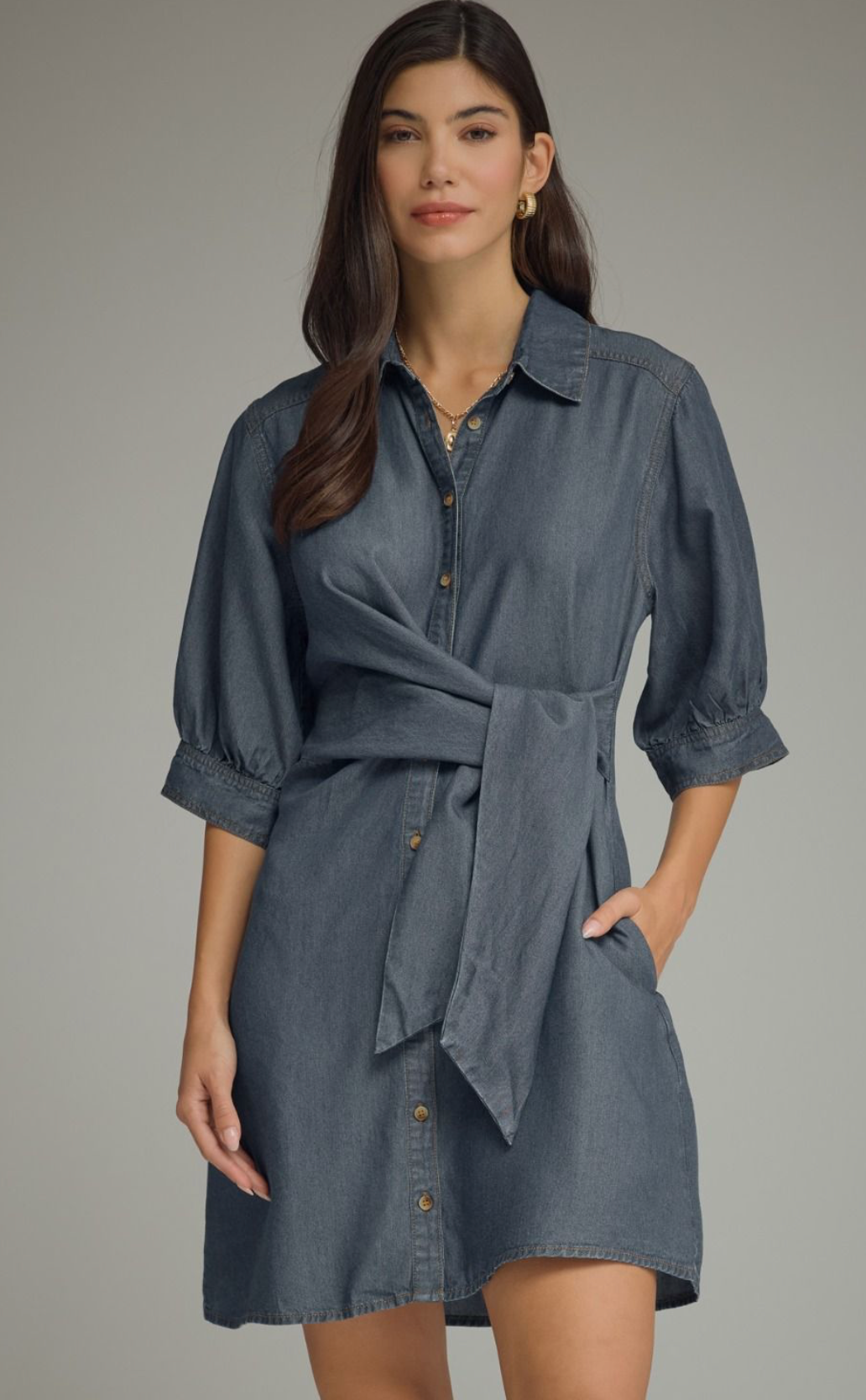 A woman modeling a blue-gray chambray shirt dress with puff sleeves, a button-down front, and a self-tie waist belt, set against a plain gray studio background.