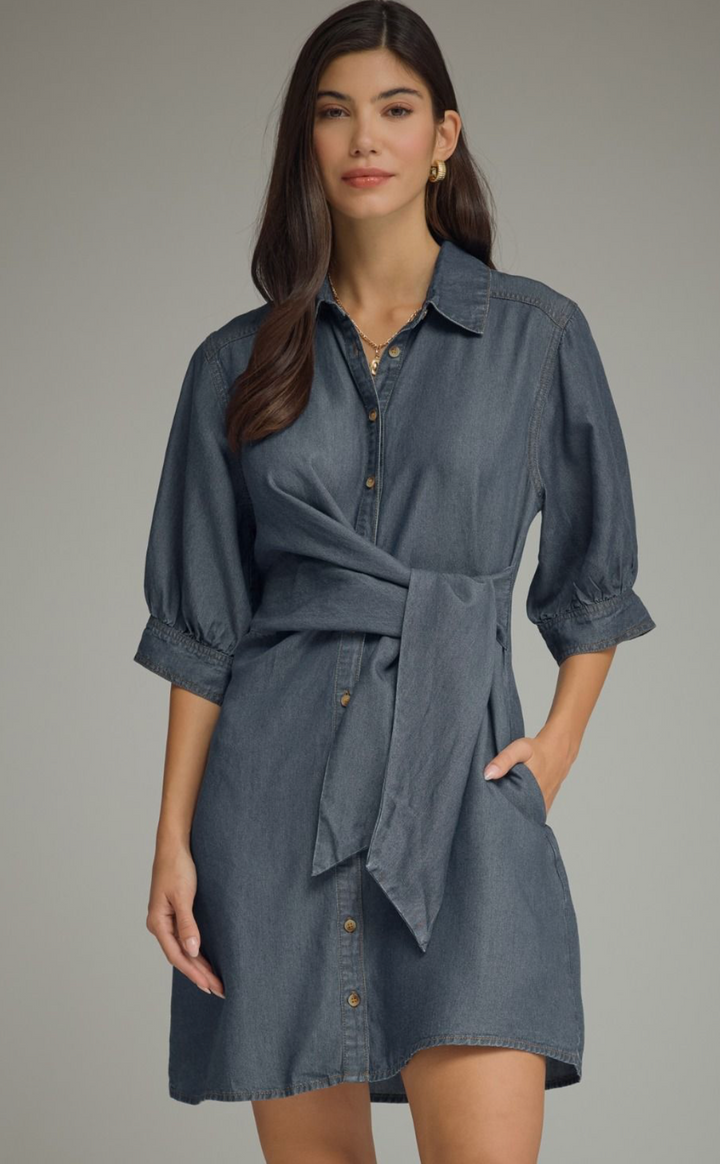 A woman modeling a blue-gray chambray shirt dress with puff sleeves, a button-down front, and a self-tie waist belt, set against a plain gray studio background.