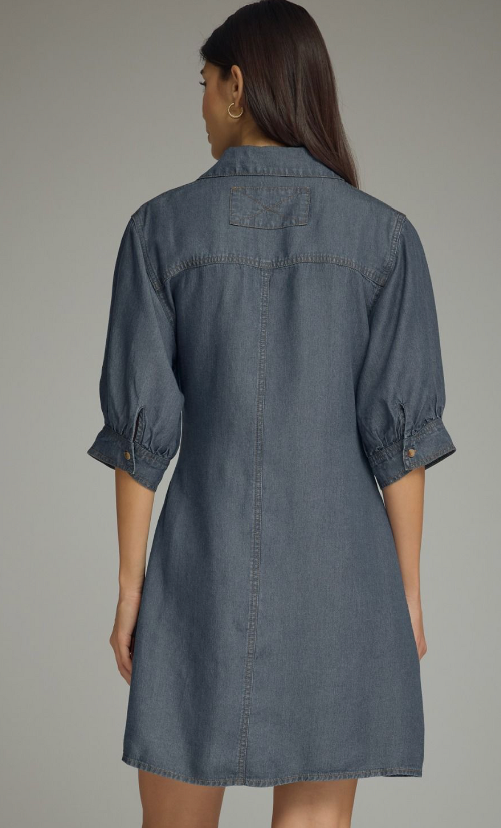 A woman shown from the back wearing a slate blue denim shirt dress with three-quarter sleeves and button cuffs, set against a plain gray studio background.