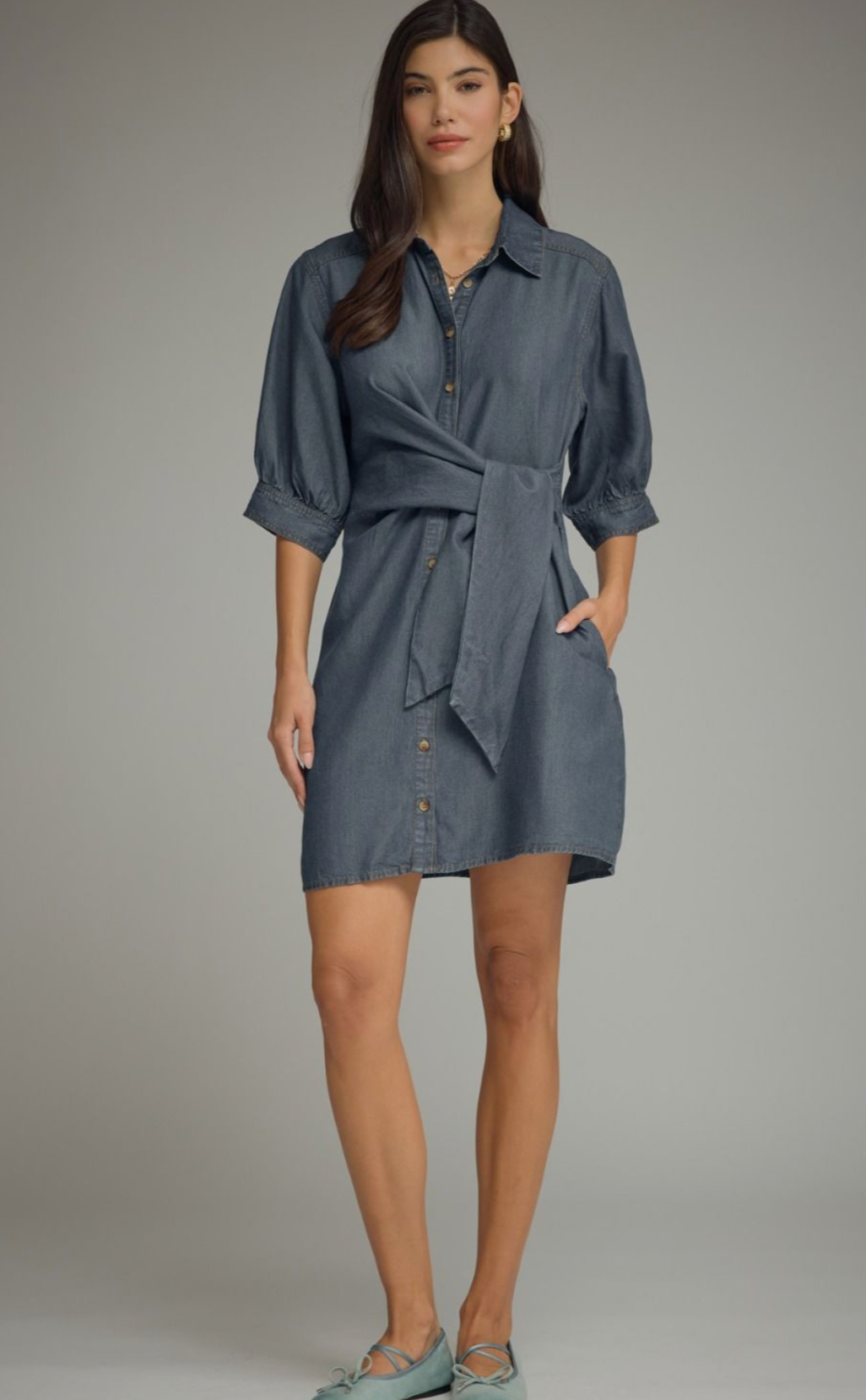 A woman modeling a slate blue button-down shirt dress with a tie waist, three-quarter sleeves, and front pockets, set against a neutral gray studio background.