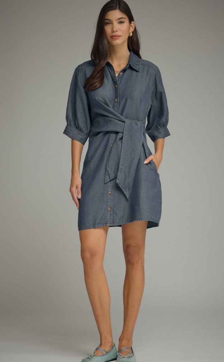 A woman modeling a slate blue button-down shirt dress with a tie waist, three-quarter sleeves, and front pockets, set against a neutral gray studio background.