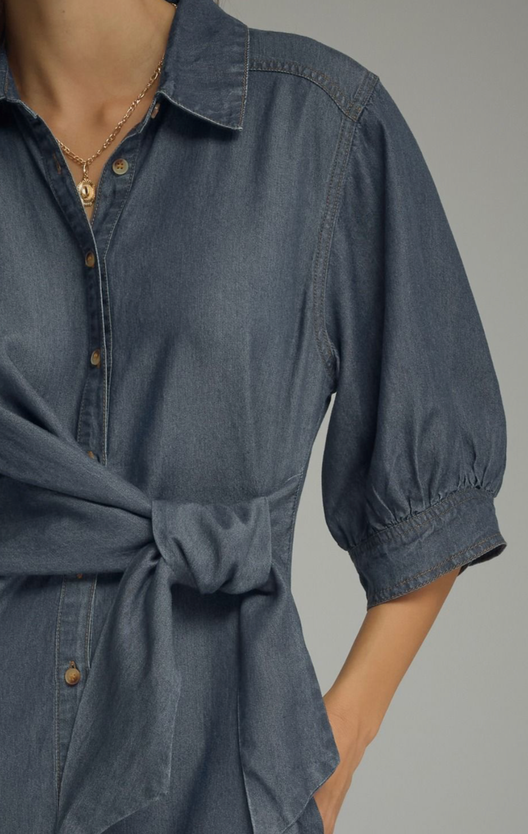 A close-up of a person wearing a dark blue denim shirt dress with puff sleeves and a tied waist, paired with a gold necklace.