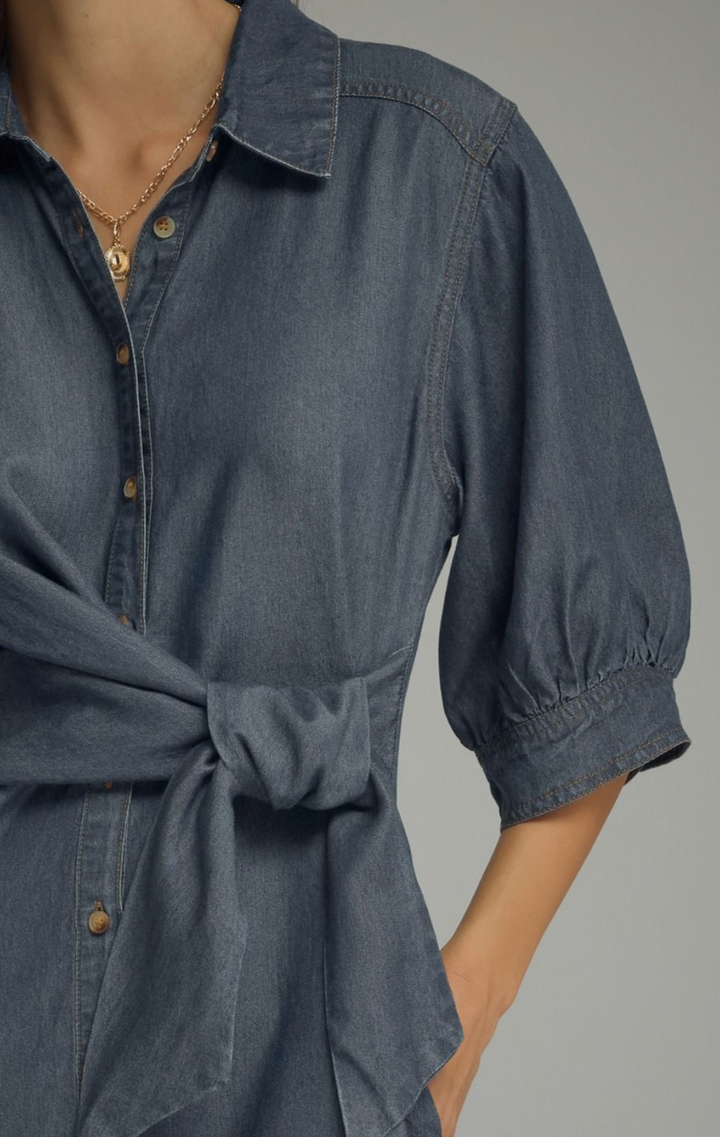 A close-up of a person wearing a dark blue denim shirt dress with puff sleeves and a tied waist, paired with a gold necklace.