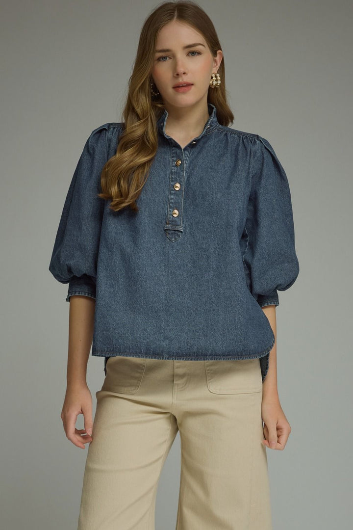 A woman wearing a blue denim blouse with puff sleeves and beige pants, standing against a plain gray studio background.
