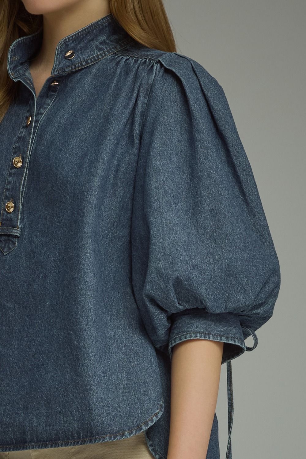 A close-up of a model wearing a blue denim blouse with voluminous puff sleeves and a buttoned collar, shown against a neutral gray background.