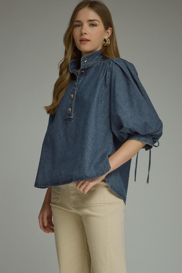 A woman modeling a blue denim blouse with voluminous puff sleeves, a high mock neck, and a buttoned chest, paired with beige pants against a neutral gray studio background.
