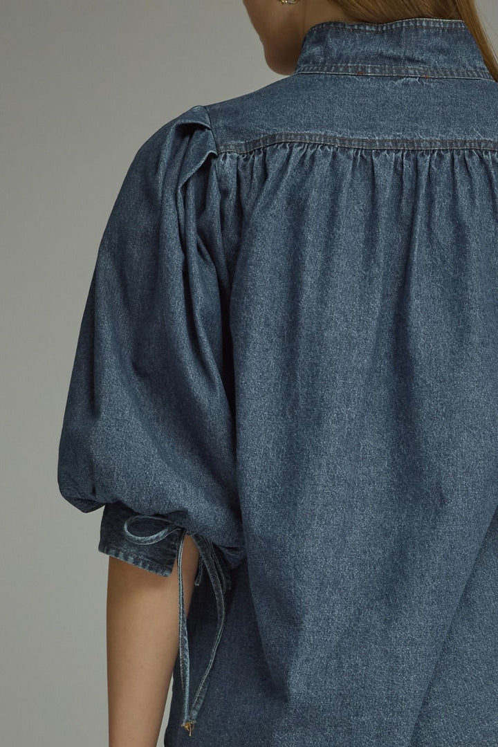 Close-up back view of a person wearing a blue denim blouse with gathered puff sleeves and side tie details at the cuffs, against a plain light gray studio background.