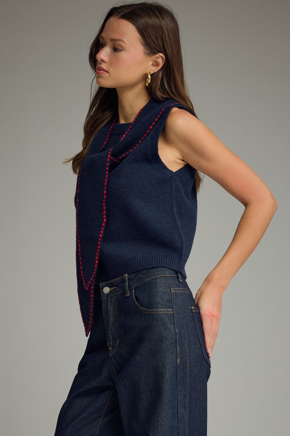 Woman wearing a navy blue sleeveless top with red trim and dark jeans on a gray background