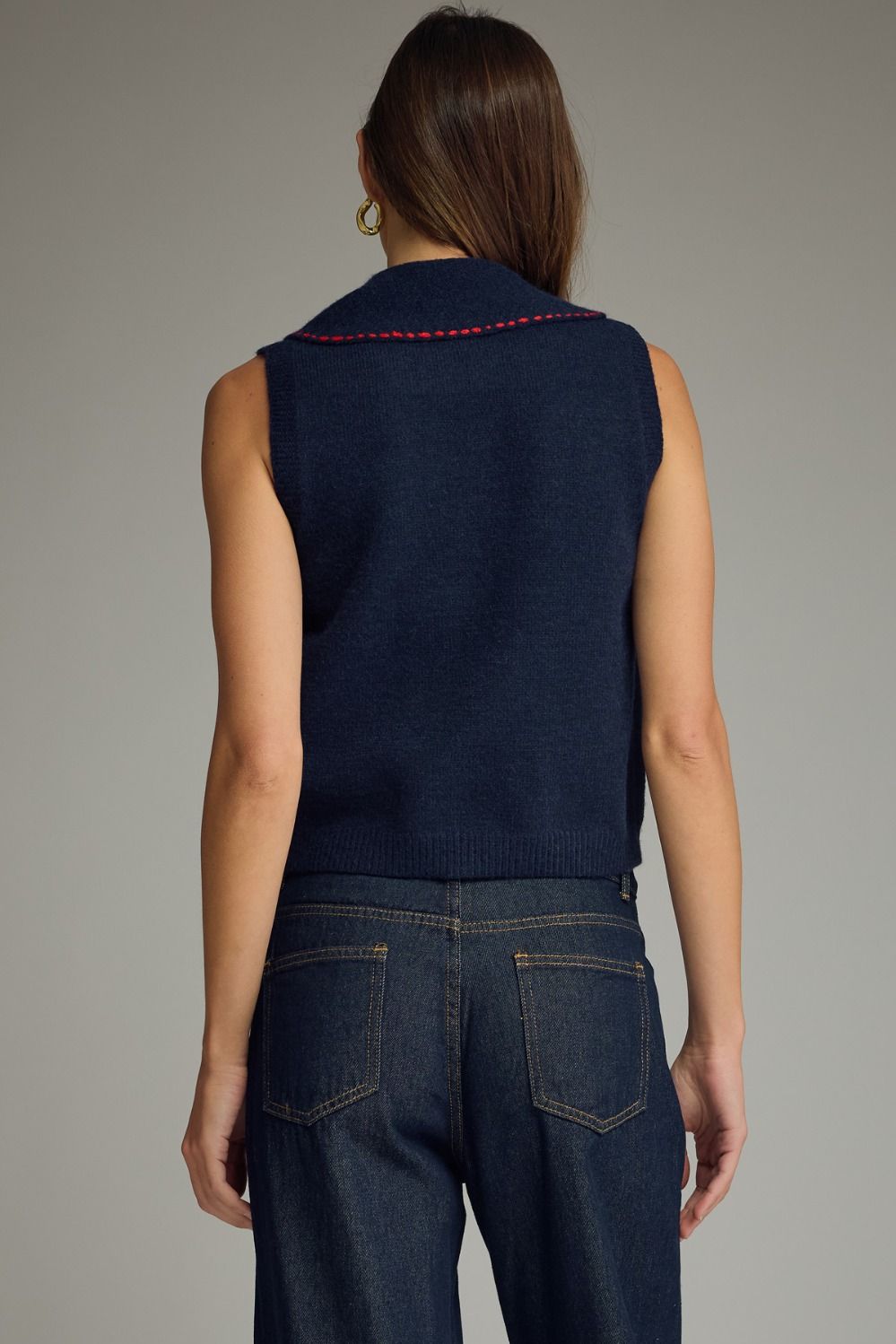 Person wearing a navy blue sleeveless top with red trim and dark jeans on a gray background