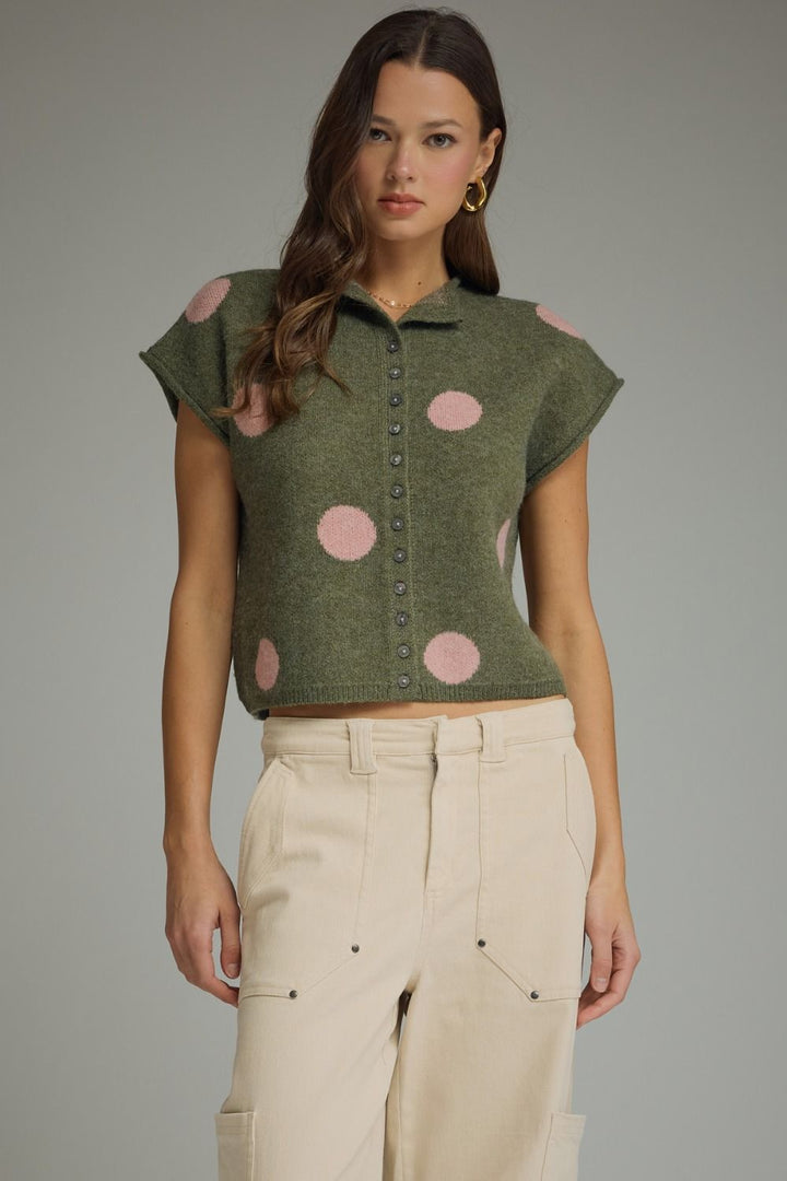 Woman wearing a green sleeveless top with pink polka dots and beige pants on a gray background