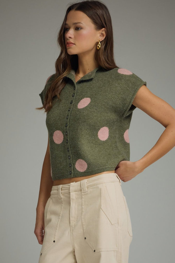 Woman wearing a green vest with pink polka dots and beige pants on a gray background