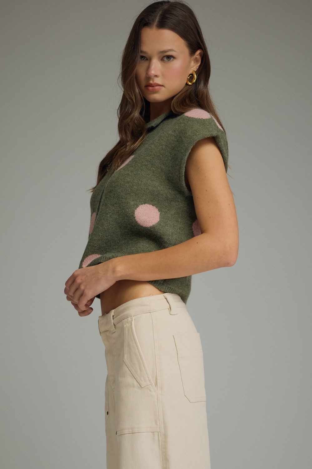 Woman wearing a green sleeveless top with pink polka dots and beige pants on a gray background