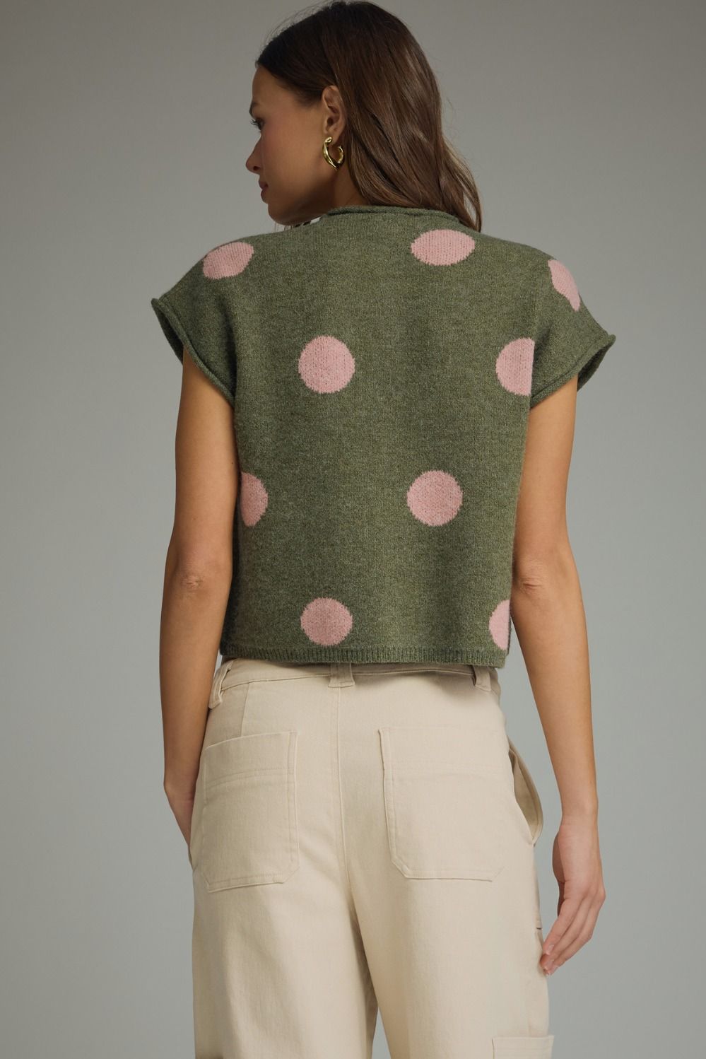 Woman wearing a green sleeveless top with pink polka dots and beige pants on a gray background