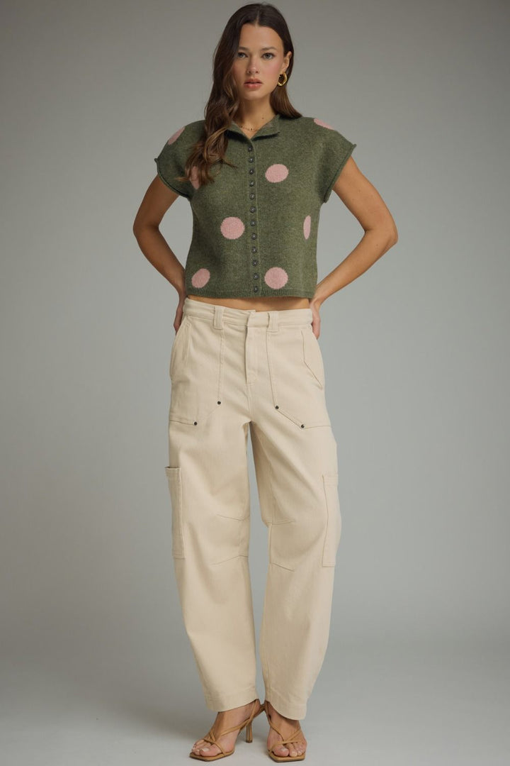 Woman wearing a green sleeveless top with pink polka dots and beige pants on a gray background