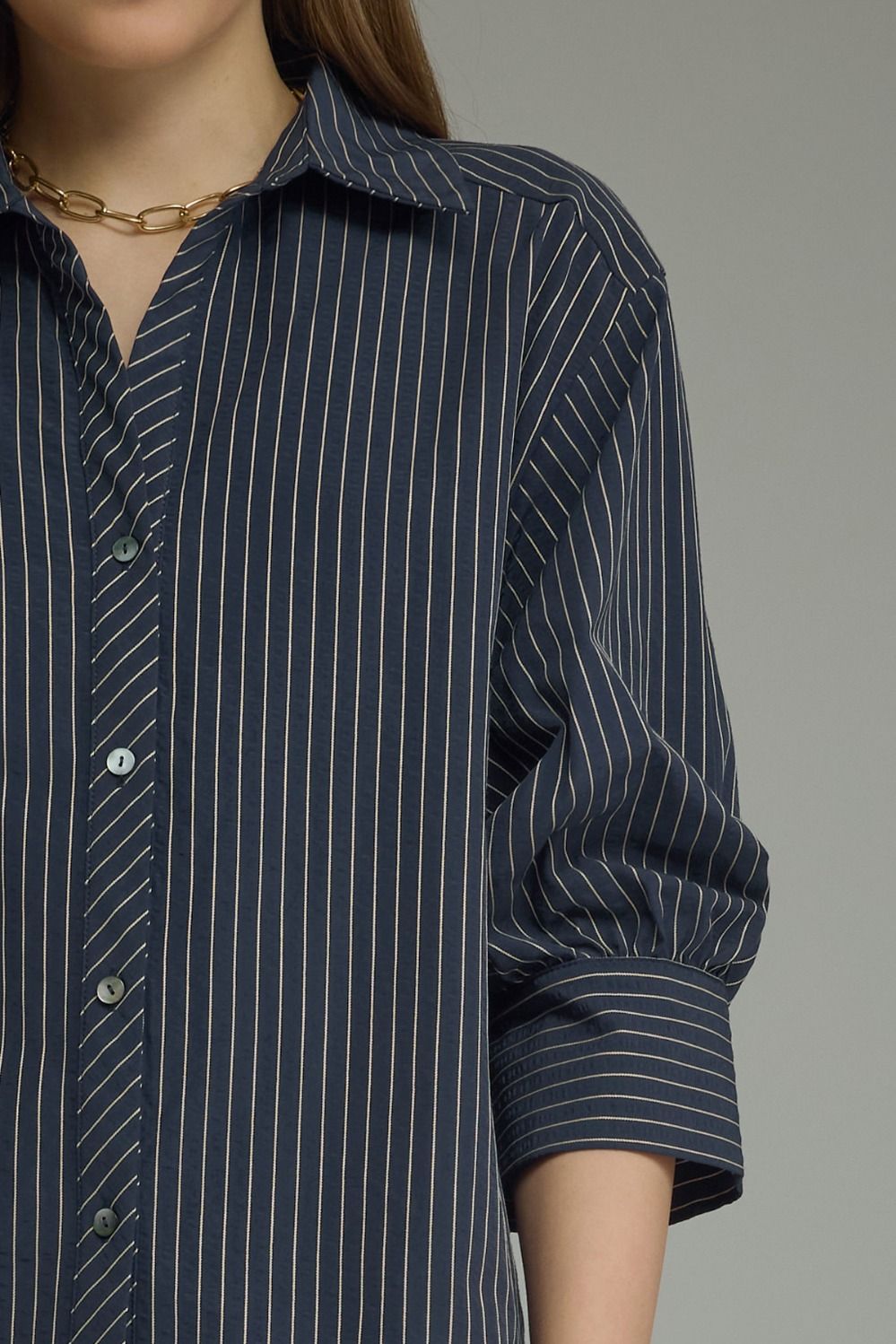 A close-up of a navy blue button-down shirt with thin white vertical pinstripes, worn by a model with a gold chain necklace.