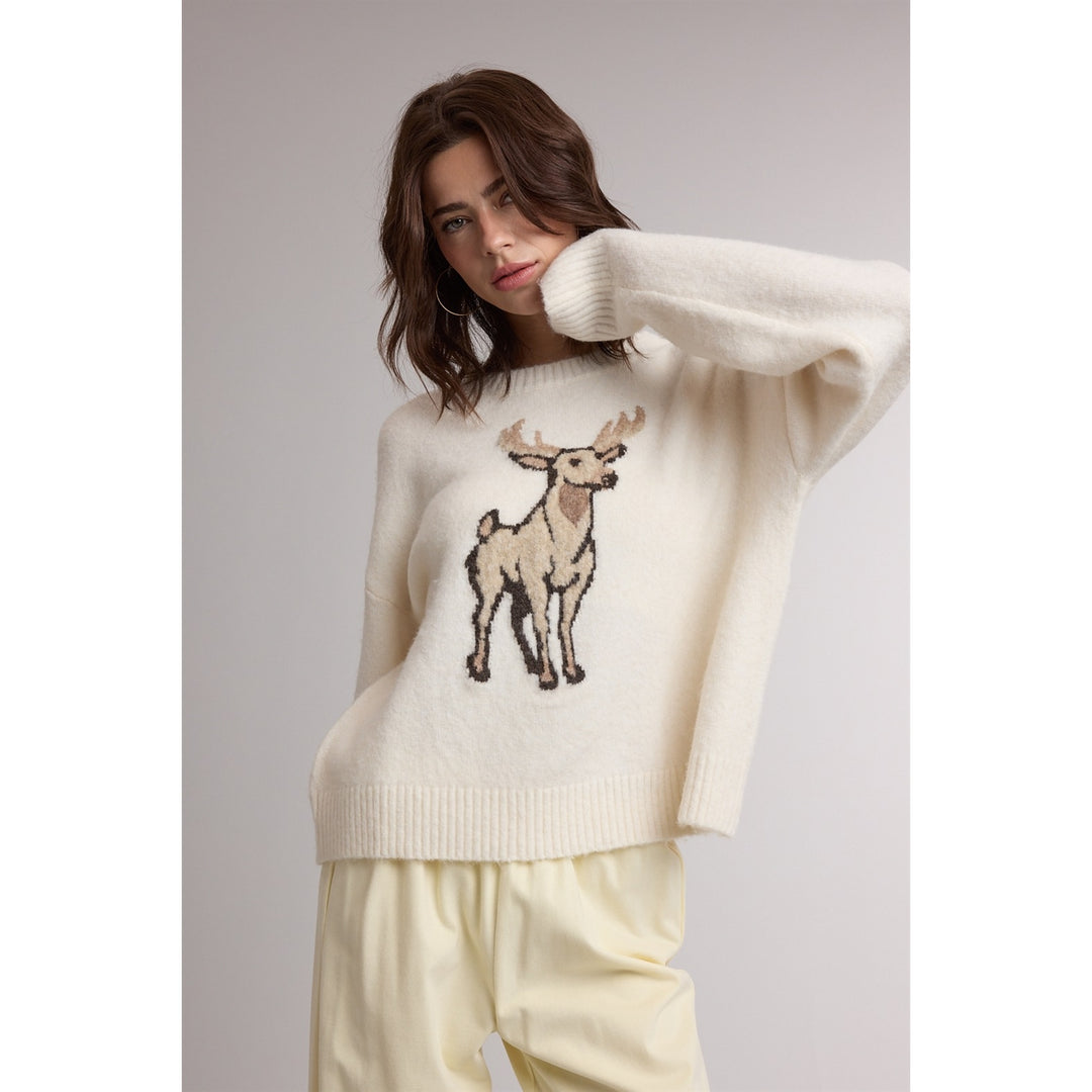 A woman wearing a cream-colored knit sweater with a detailed embroidered deer motif on the front, paired with matching cream pants, set against a plain light gray studio background.